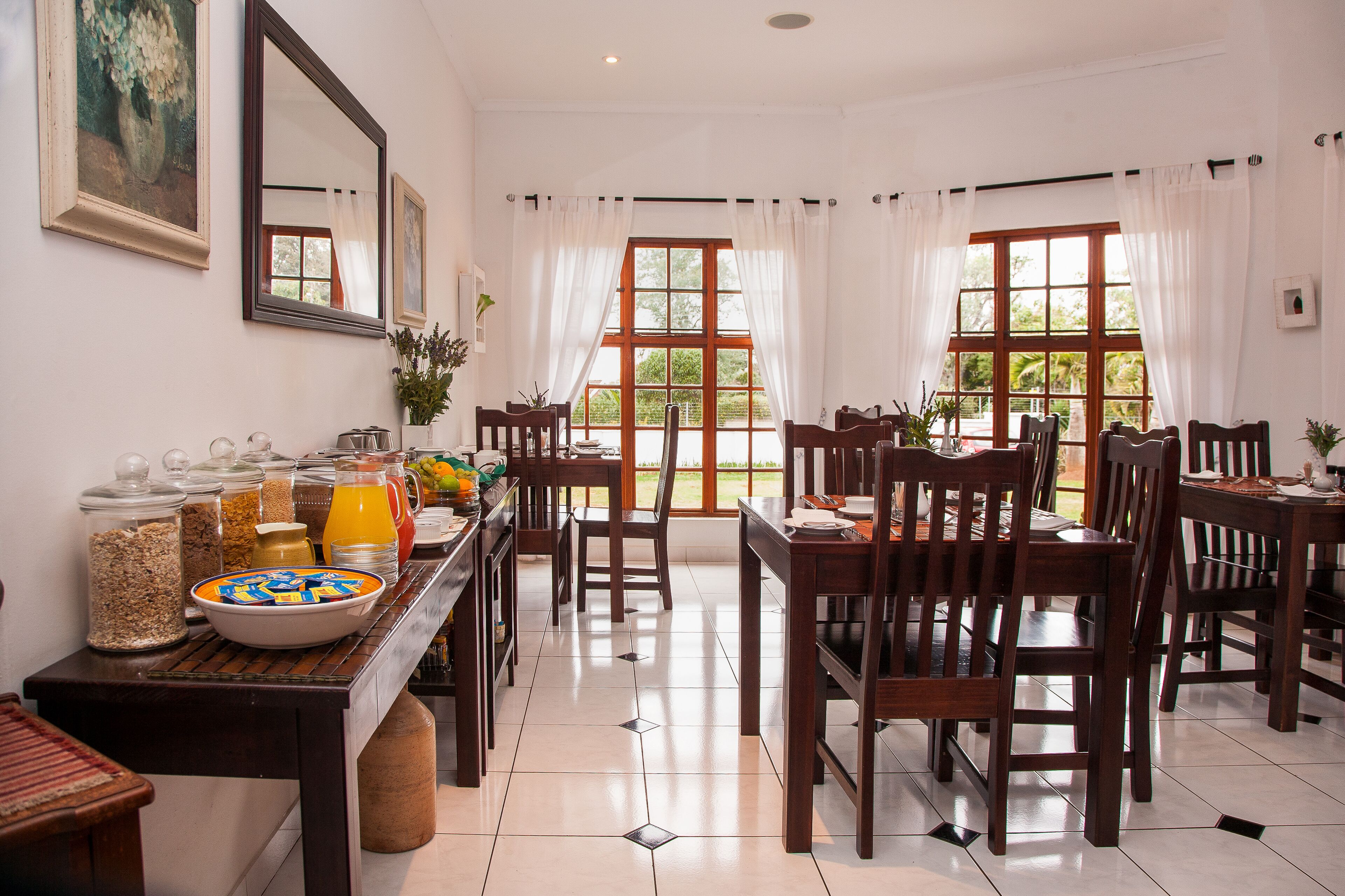 A dining area with wooden tables and chairs, a buffet with food and drinks, and large windows with curtains.