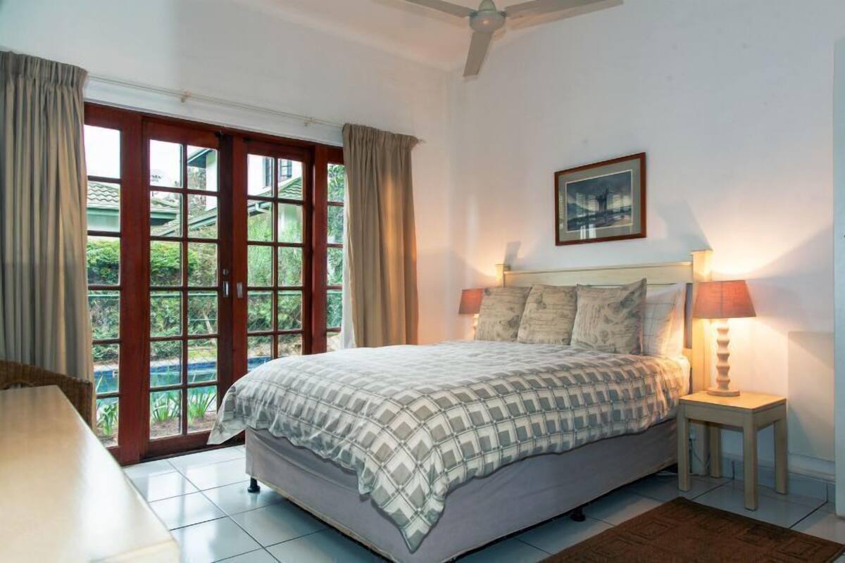 A bedroom with a bed, a nightstand, a ceiling fan, a window with curtains, and a framed picture on the wall.