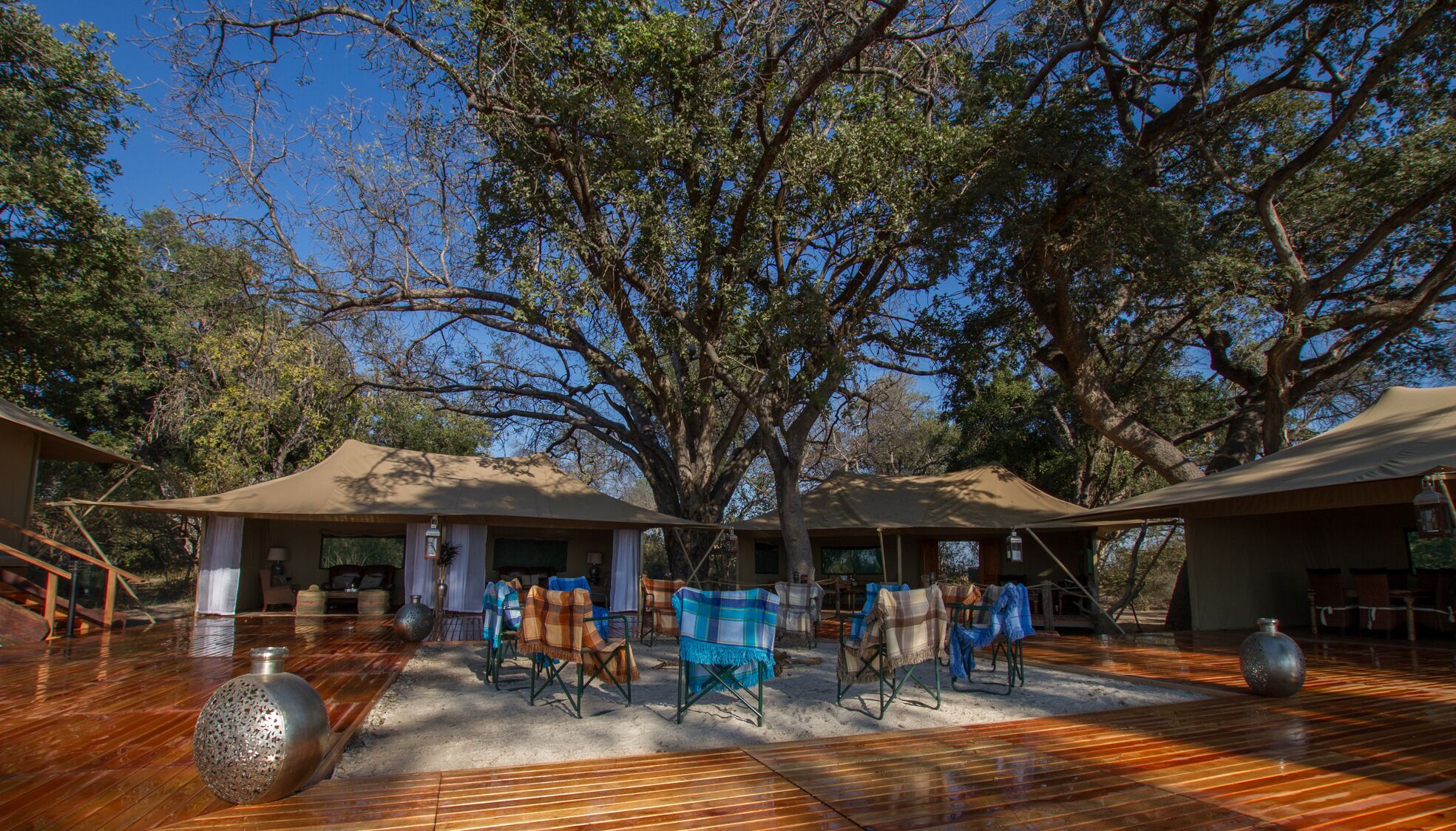 A covered outdoor area with wooden flooring, seating, and a large tree providing shade.