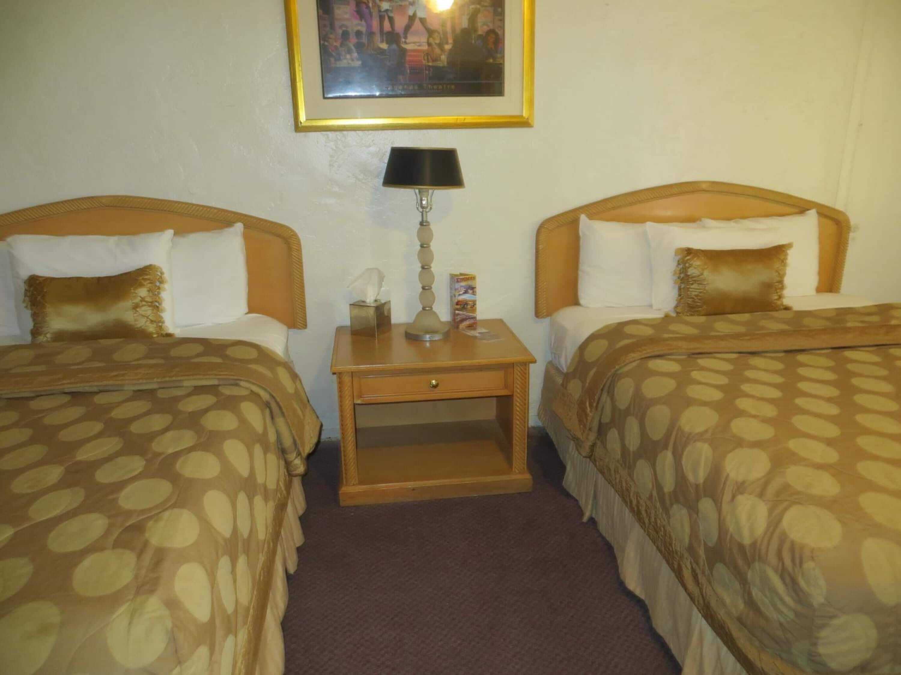 A hotel room with two beds, each with a headboard, a bedside table with a lamp, and a framed picture on the wall.