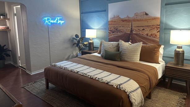 A bedroom with a bed, bedside lamps, a neon sign, and a large landscape painting on the wall. - El Trovatore Motel (Kingman)