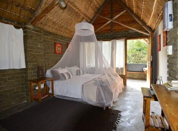 A bedroom with a bed under a mosquito net, a wooden nightstand, and a stone wall.
