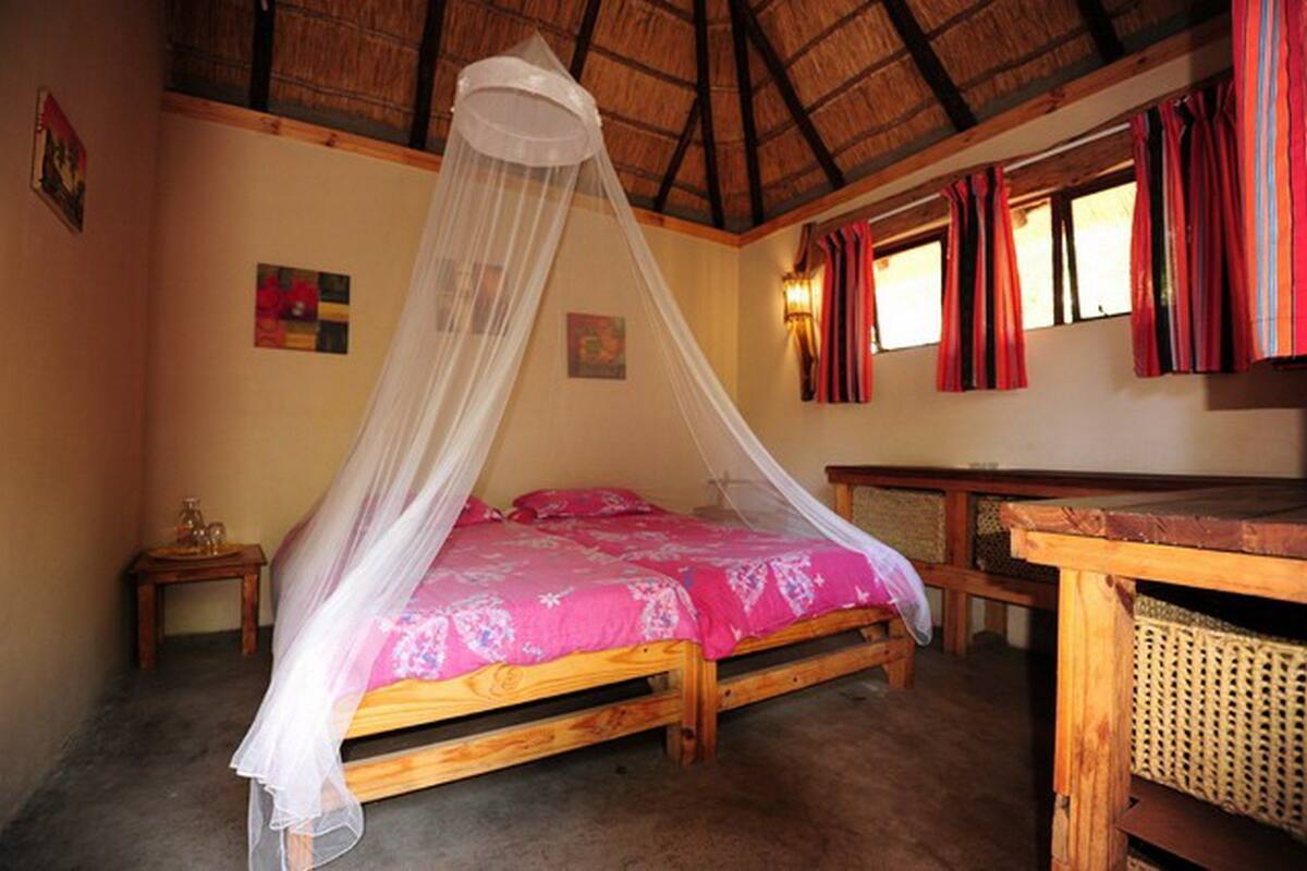 A room with a bed under a mosquito net, wooden furniture, and a thatched roof.