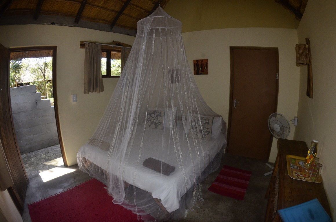 Un dormitorio con cama cubierta por una mosquitera, una puerta de madera, un ventilador y una mesita con una botella y vasos.