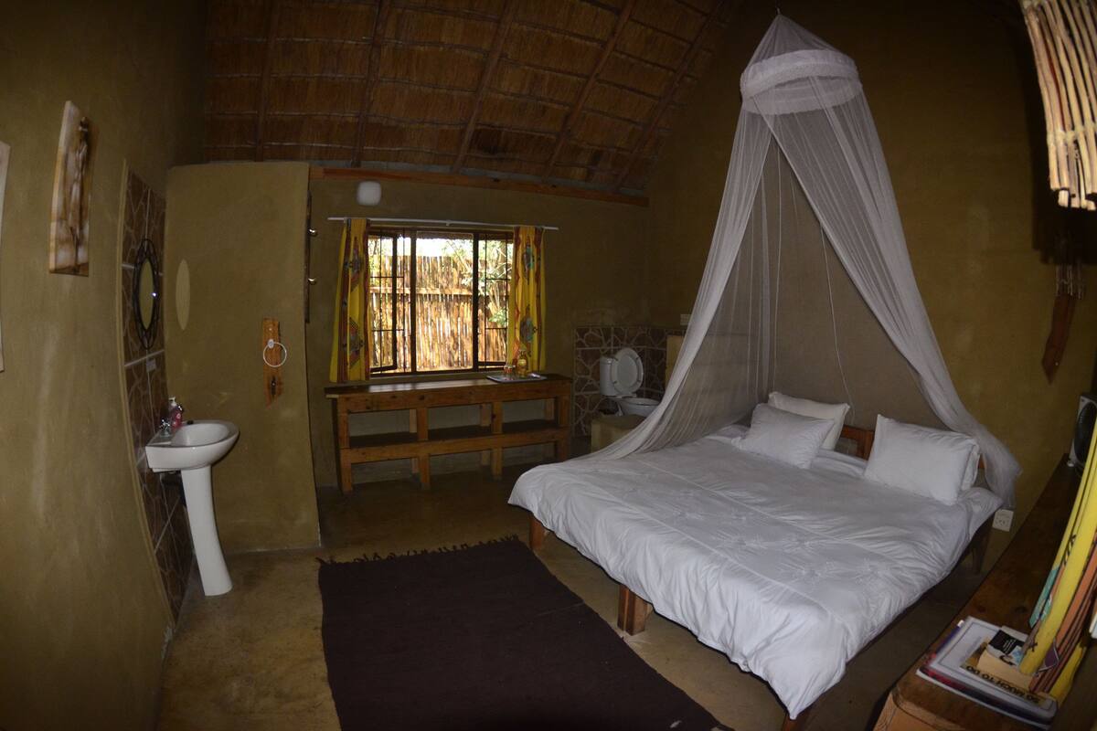 A bedroom with a bed, a bedside table, a window with curtains, a sink, and a mosquito net over the bed.