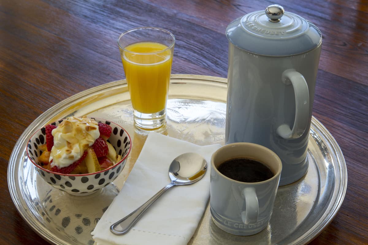 A tray with a polka-dotted bowl of fruit, a glass of orange juice, a coffee pot, a cup of coffee, and a spoon.