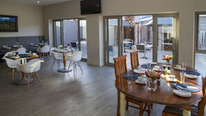 A dining area with a wooden table set for four, surrounded by white chairs. There is a television mounted on the wall above the seating area. - Ocean Bay Guest House (Gqeberha (Port Elizabeth))