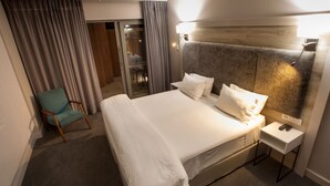 A neatly made bed with white linens, a bedside table with a remote control, and a chair with a teal cushion. - Ocean Bay Guest House (Gqeberha (Port Elizabeth))