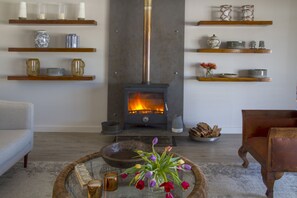 A living room with a wood-burning stove, shelves with decorative items, a sofa, and a coffee table with flowers. - Ocean Bay Guest House (Gqeberha (Port Elizabeth))