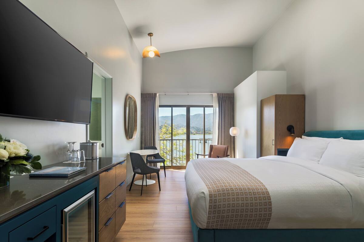A modern hotel room with a large bed, a dining area, and a view of a lake.