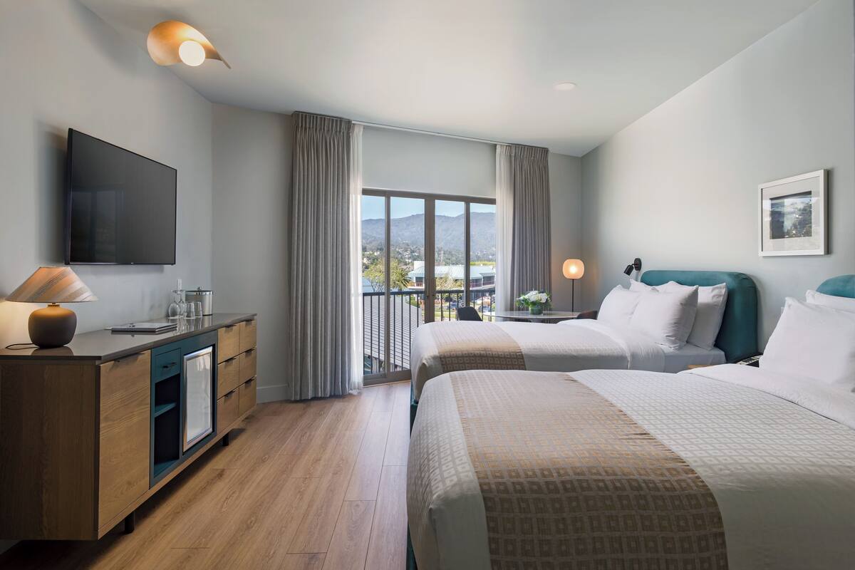 A hotel room with two beds, a desk with a lamp, a flat-screen TV, and a view of the mountains.