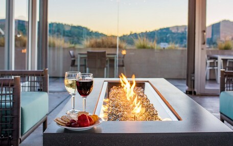 A fire feature with a view of mountains, a table with a plate of snacks and two glasses of wine, and wicker chairs.