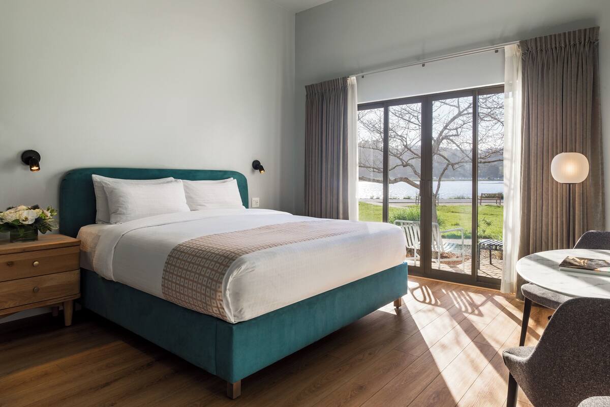 A bedroom with a large bed, a nightstand, a lamp, and a view of a park through a glass door.
