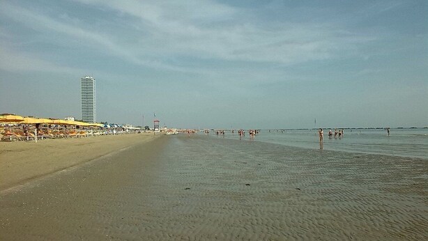 A beach with a long stretch of sand, a few umbrellas, and a tall building in the background. - Jole (Cesenatico)