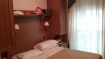 Ein Hotelzimmer mit Bett, Nachttisch, Lampe, Kleiderschrank und einem Fenster mit Vorhängen.