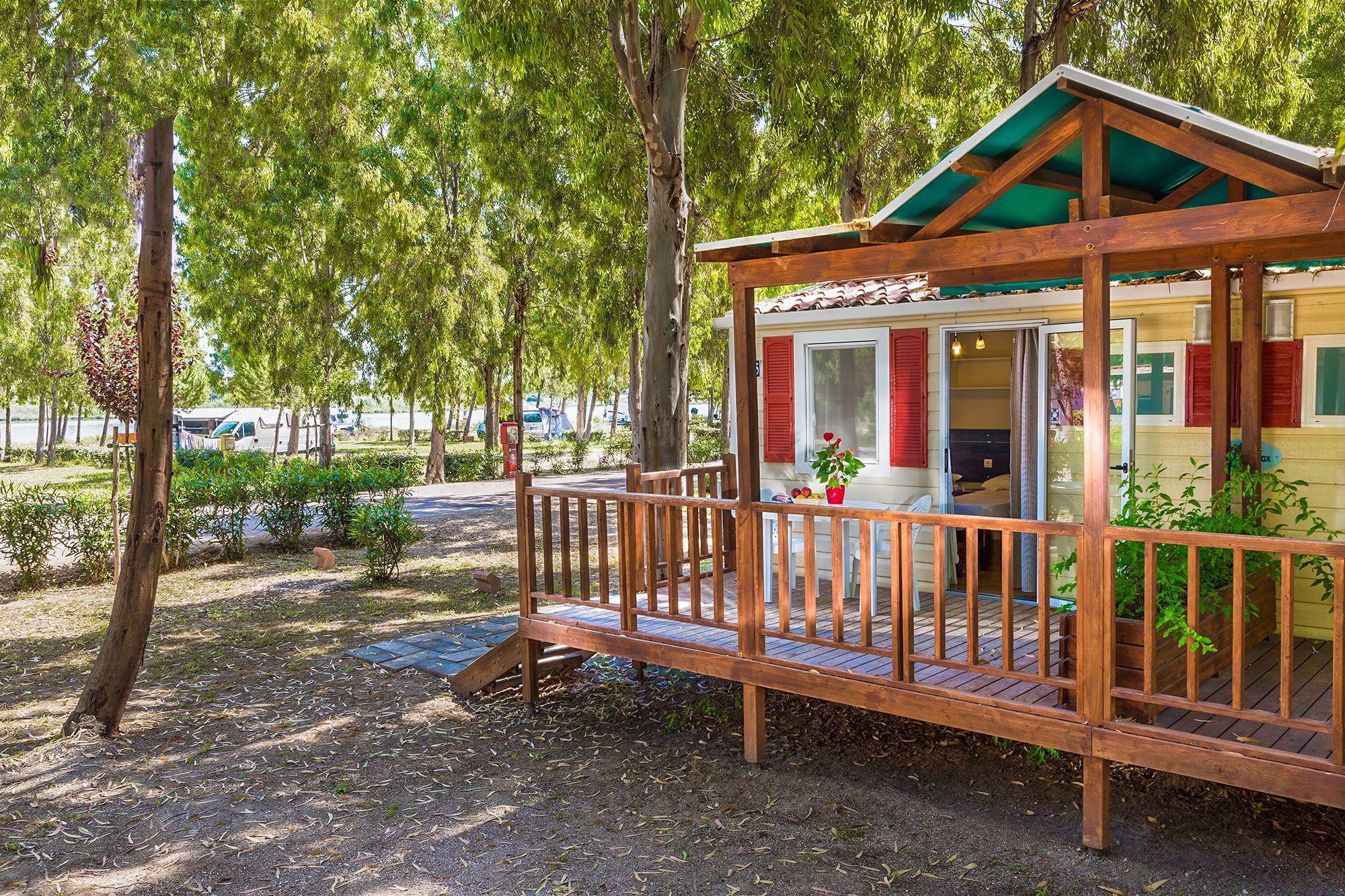 A wooden cabin with a balcony, surrounded by trees and a gravel path.