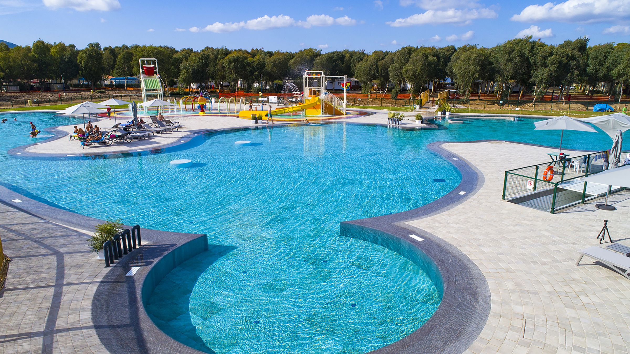 A large outdoor swimming pool with multiple lanes and a water slide, surrounded by lounge chairs and umbrellas.