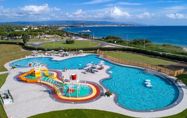 A large outdoor swimming pool with multiple lanes and a water slide, surrounded by a grassy area and a view of the sea. - Camping Village Laguna Blu (Alghero)