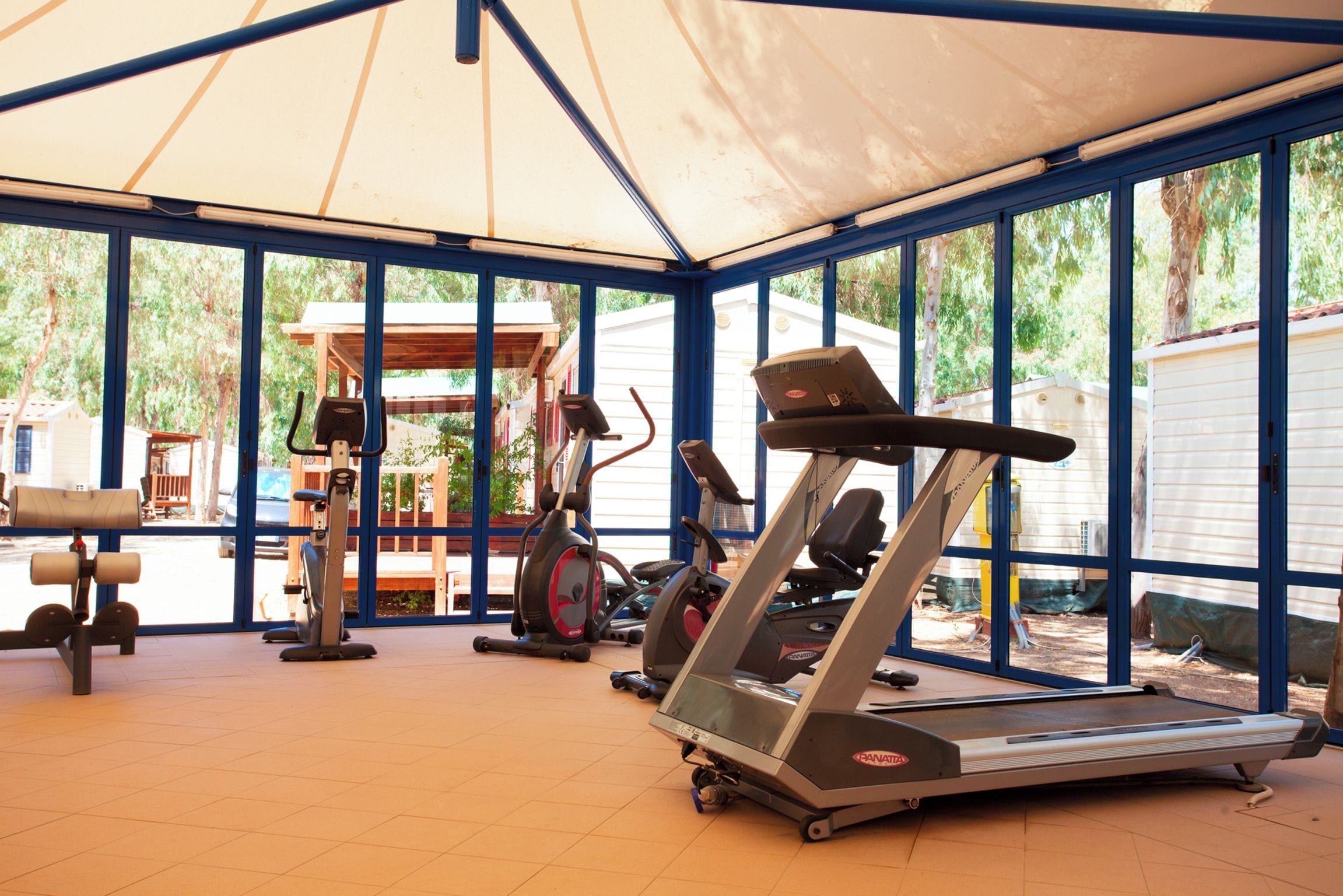 Un'area fitness all'aperto coperta, con attrezzature da palestra, tra cui tapis roulant e cyclette, sotto una grande tettoia.