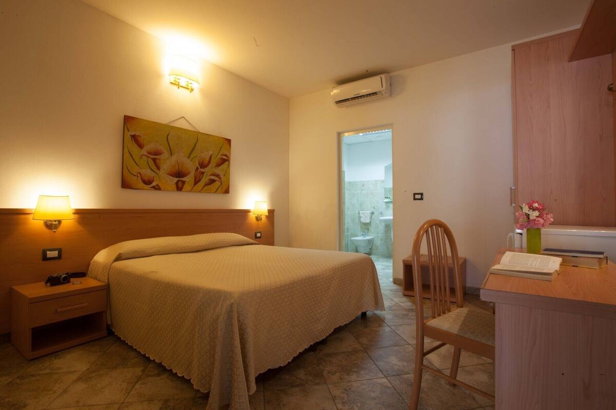 A hotel room with a bed, bedside lamps, a desk with a chair, a painting on the wall, and an air conditioning unit.