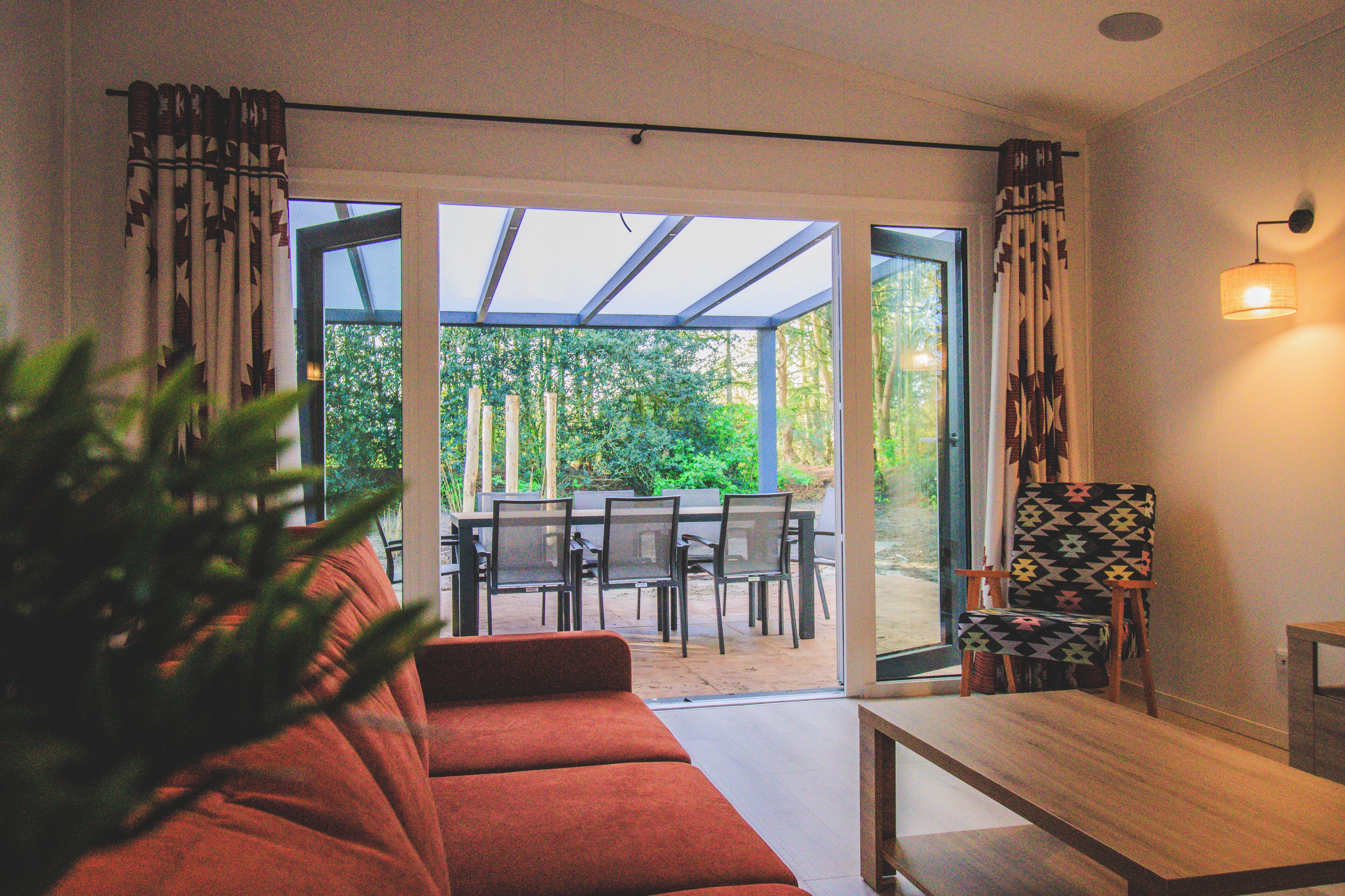 A modern living room with a sofa, a wooden coffee table, and a patterned armchair. There is a glass door leading to an outdoor patio with a table and chairs.