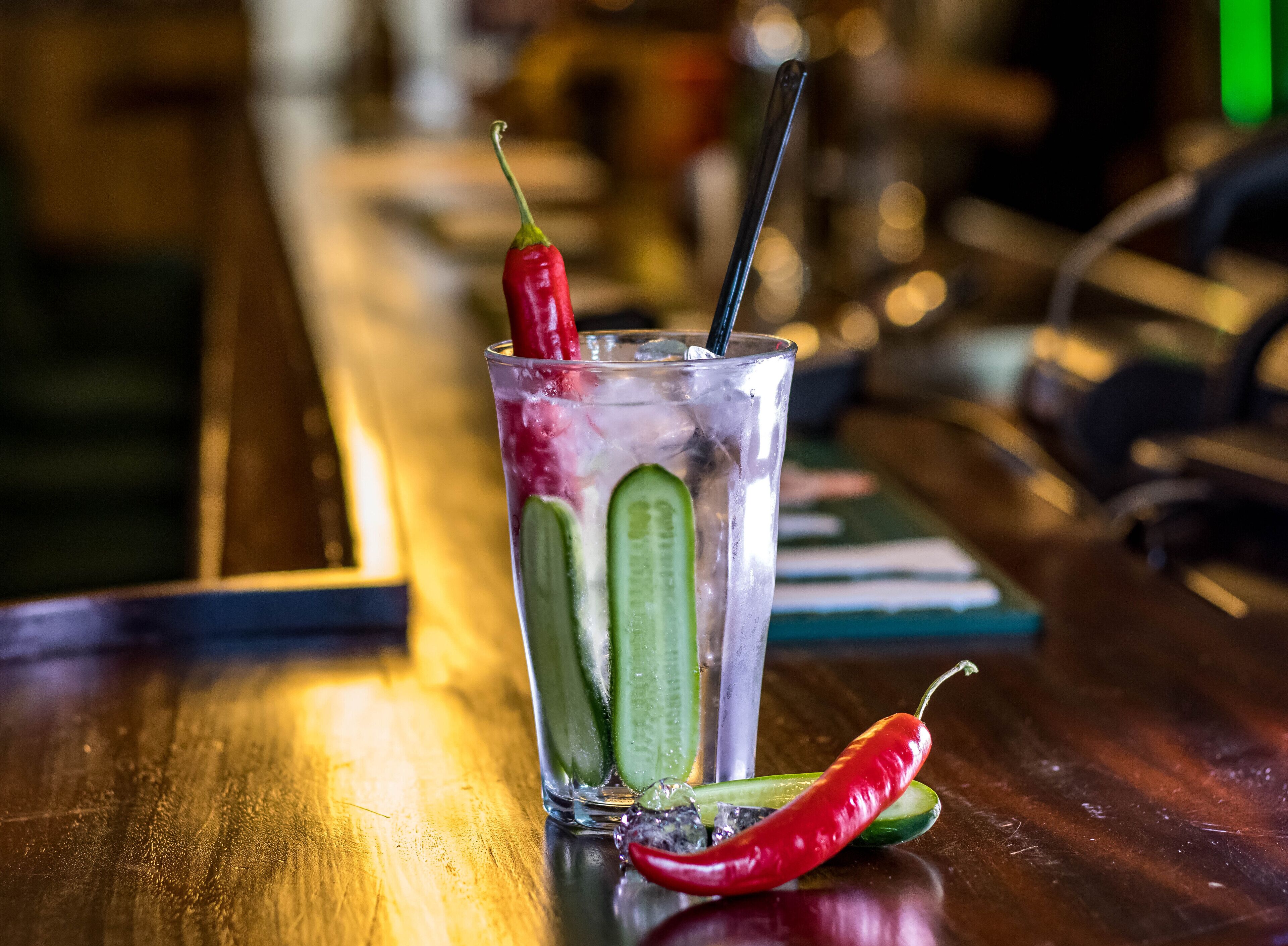Un verre transparent rempli de glaçons, un piment rouge et une tranche de concombre, le tout posé sur le comptoir en bois d’un bar.