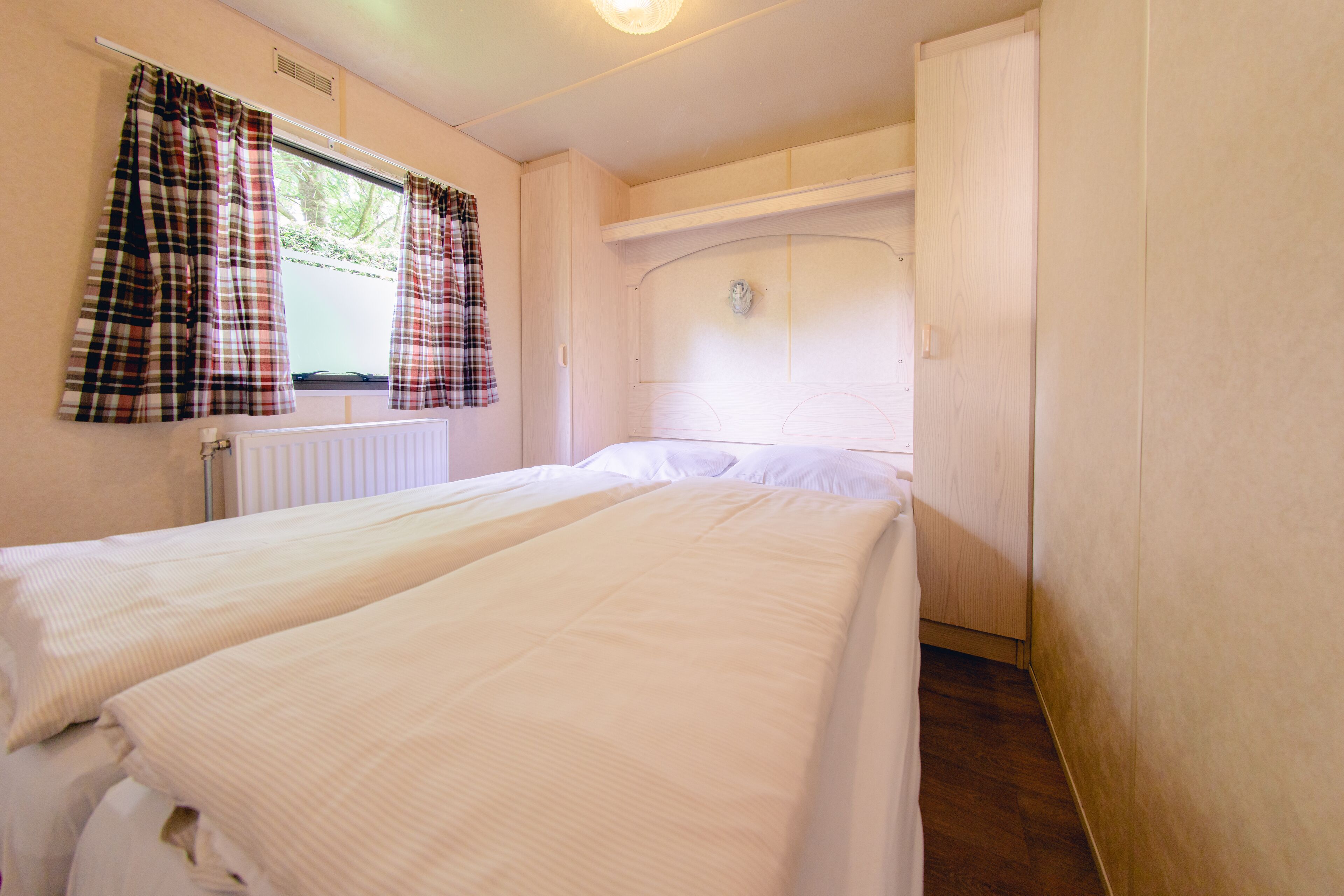 A bedroom with two beds, a window with plaid curtains, a radiator, and a wooden door.