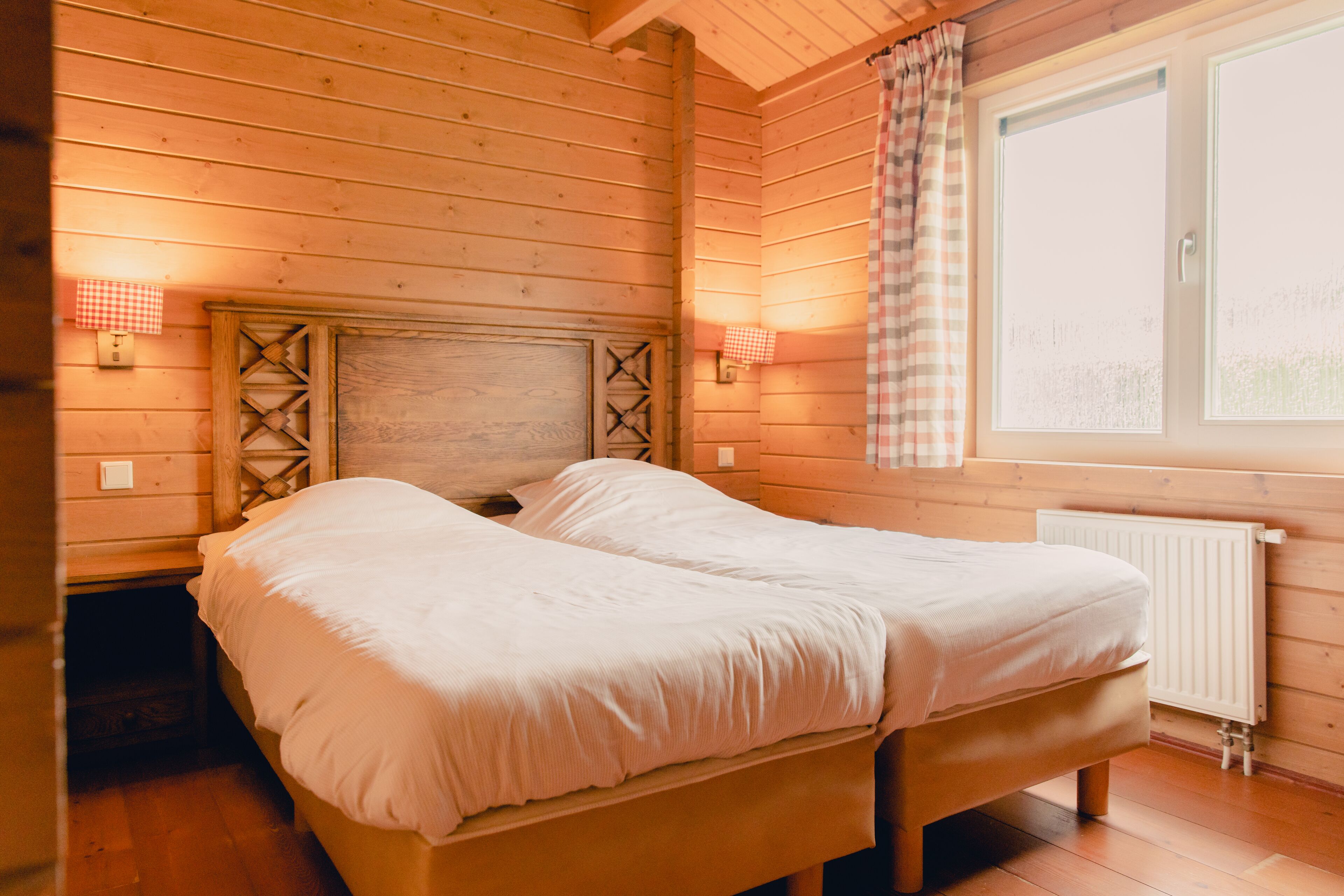 A wooden cabin room with a bed, two bedside lamps, and a window with a checkered curtain.