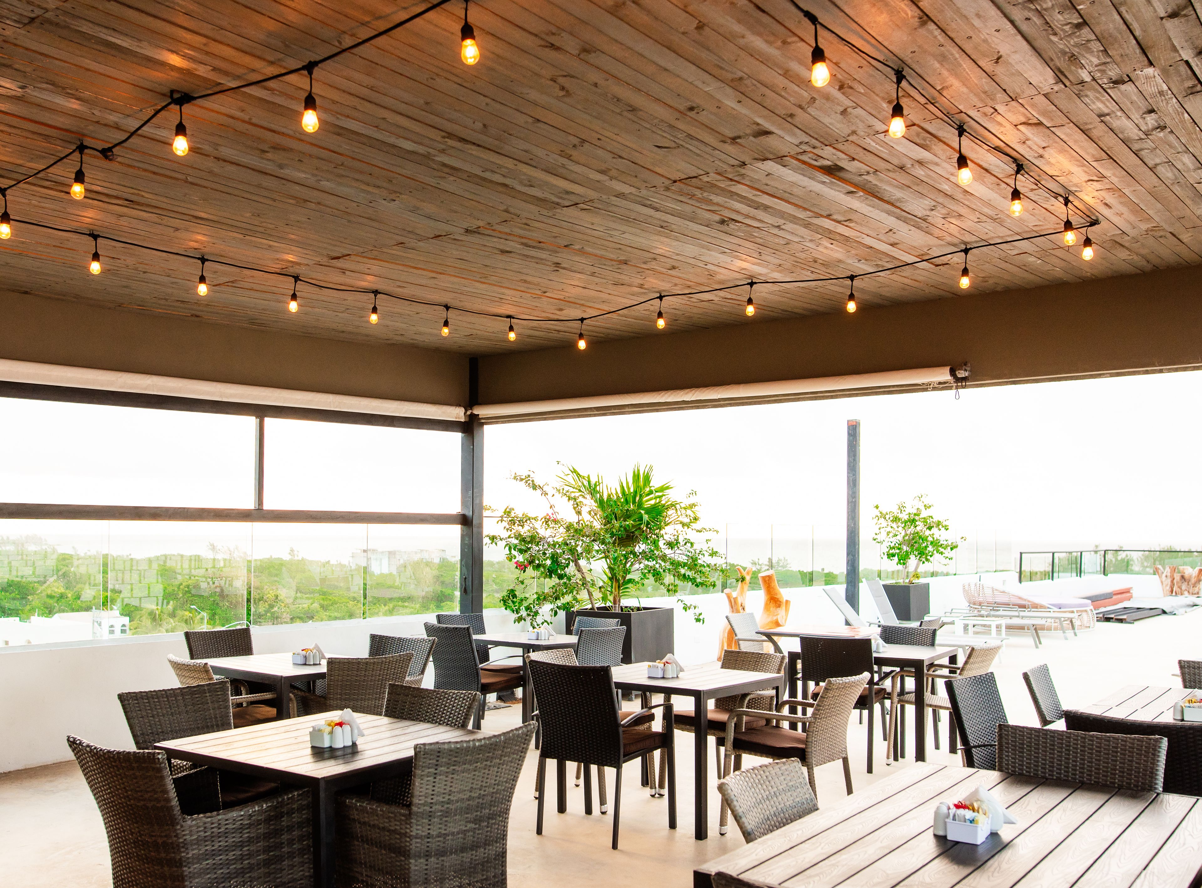 An outdoor dining area with wicker furniture, string lights, and large windows offering a view of greenery and buildings.