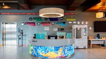 Zona de recepción de hotel con diseño moderno, que incluye un mostrador de recepción, un reloj digital y una pared con nombres de ciudades en varios idiomas.