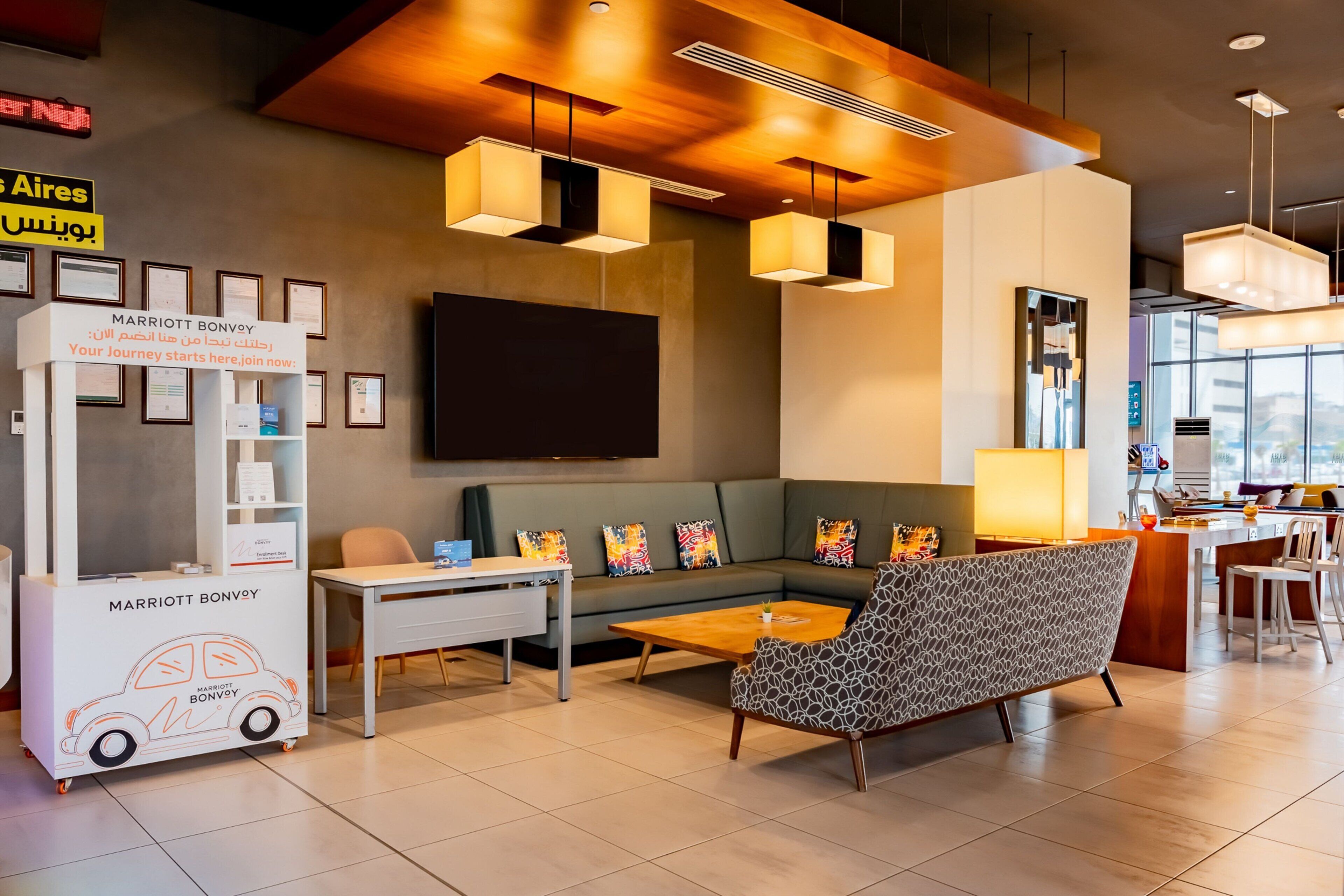 A modern hotel lobby with a seating area, a reception desk, and a large flat-screen TV mounted on the wall.