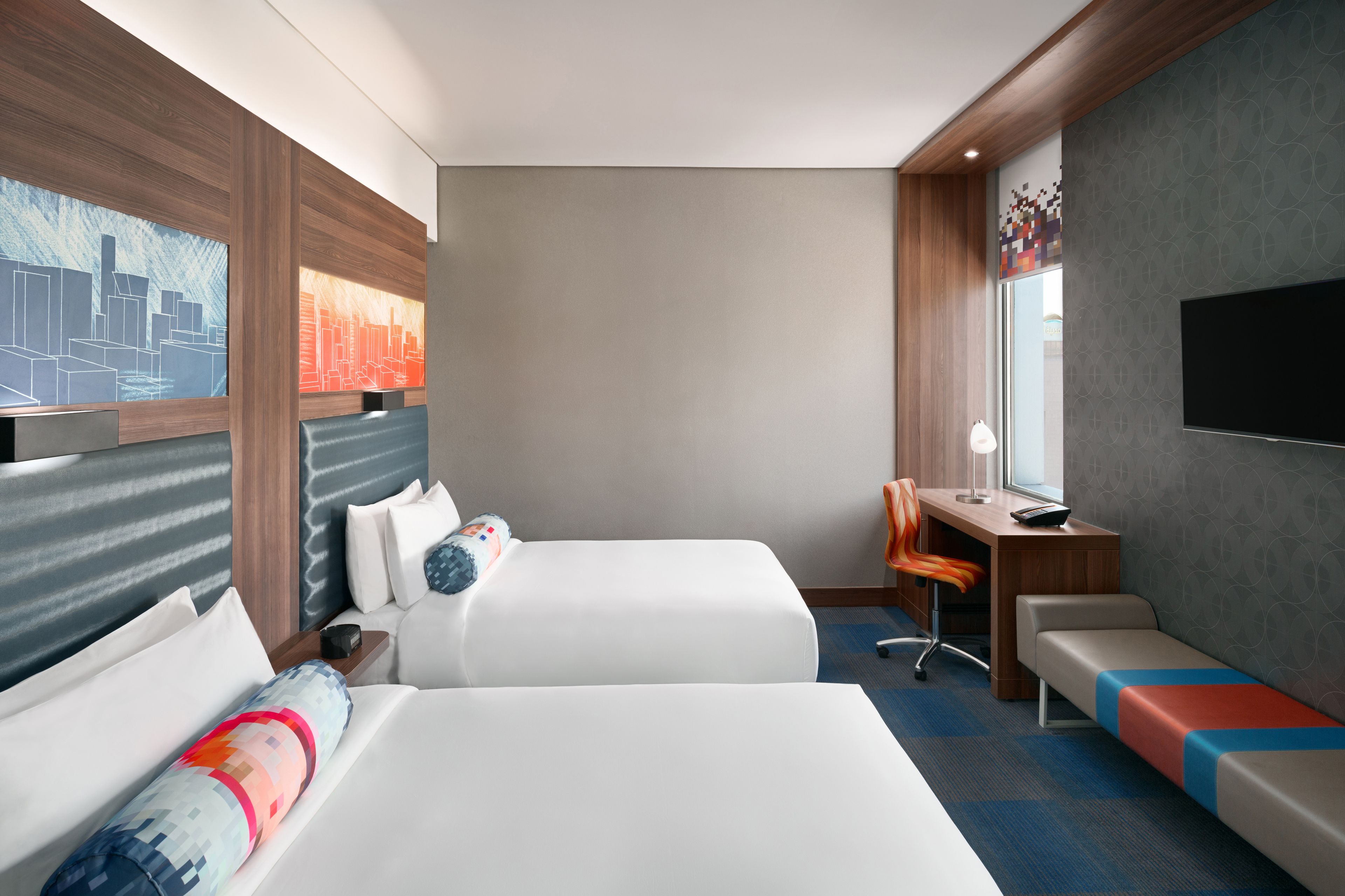 Habitación de hotel con dos camas, un escritorio, una silla, un banco, un televisor y obras de arte en las paredes.