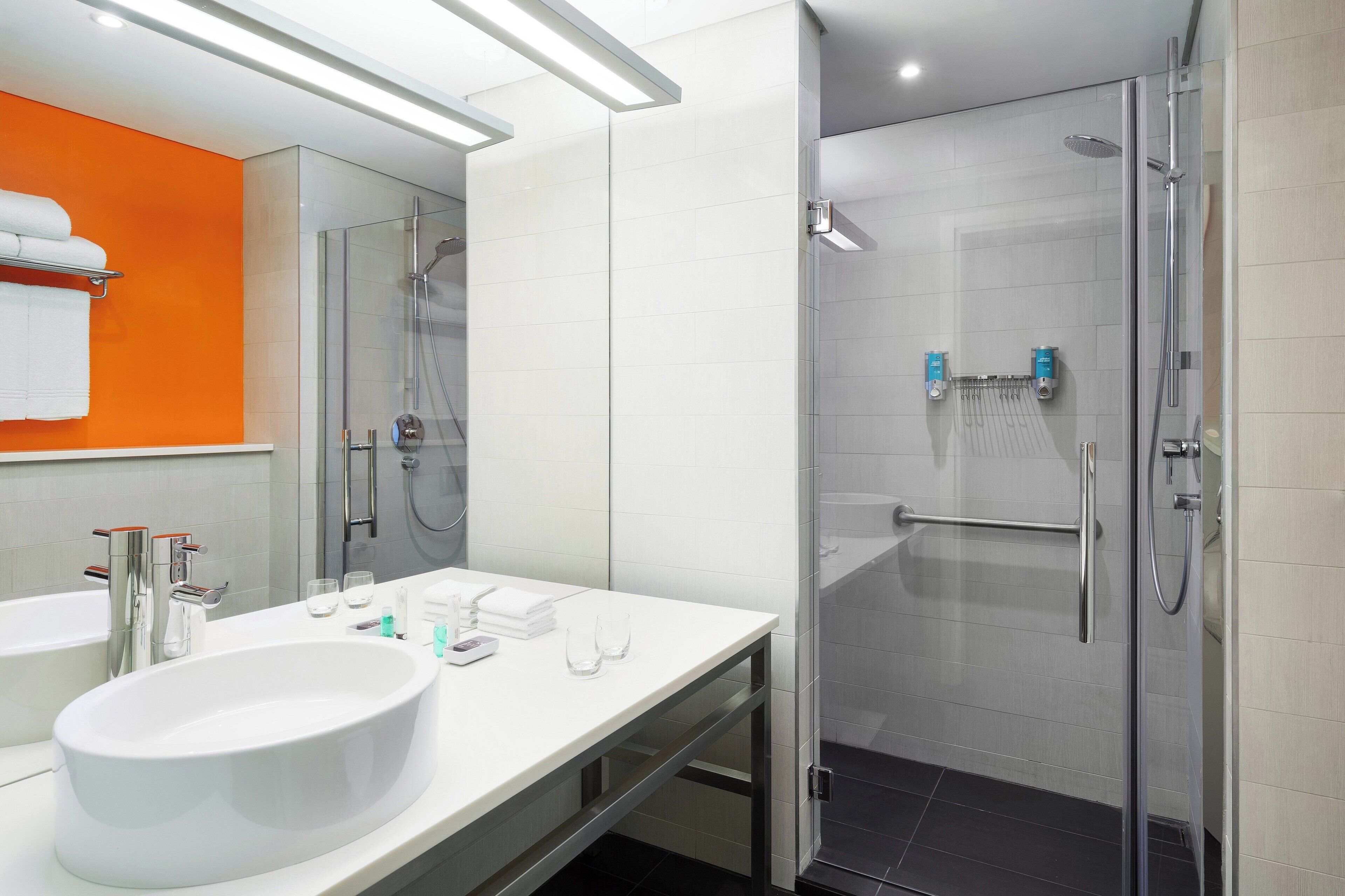 Un baño moderno con un lavamanos blanco grande, dos espejos, cabina de ducha de vidrio y un perchero para toallas.