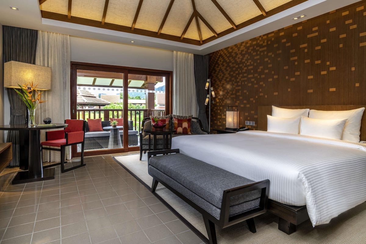 A hotel room with a large bed, a desk with a chair, a bench, and a view of the outdoors.