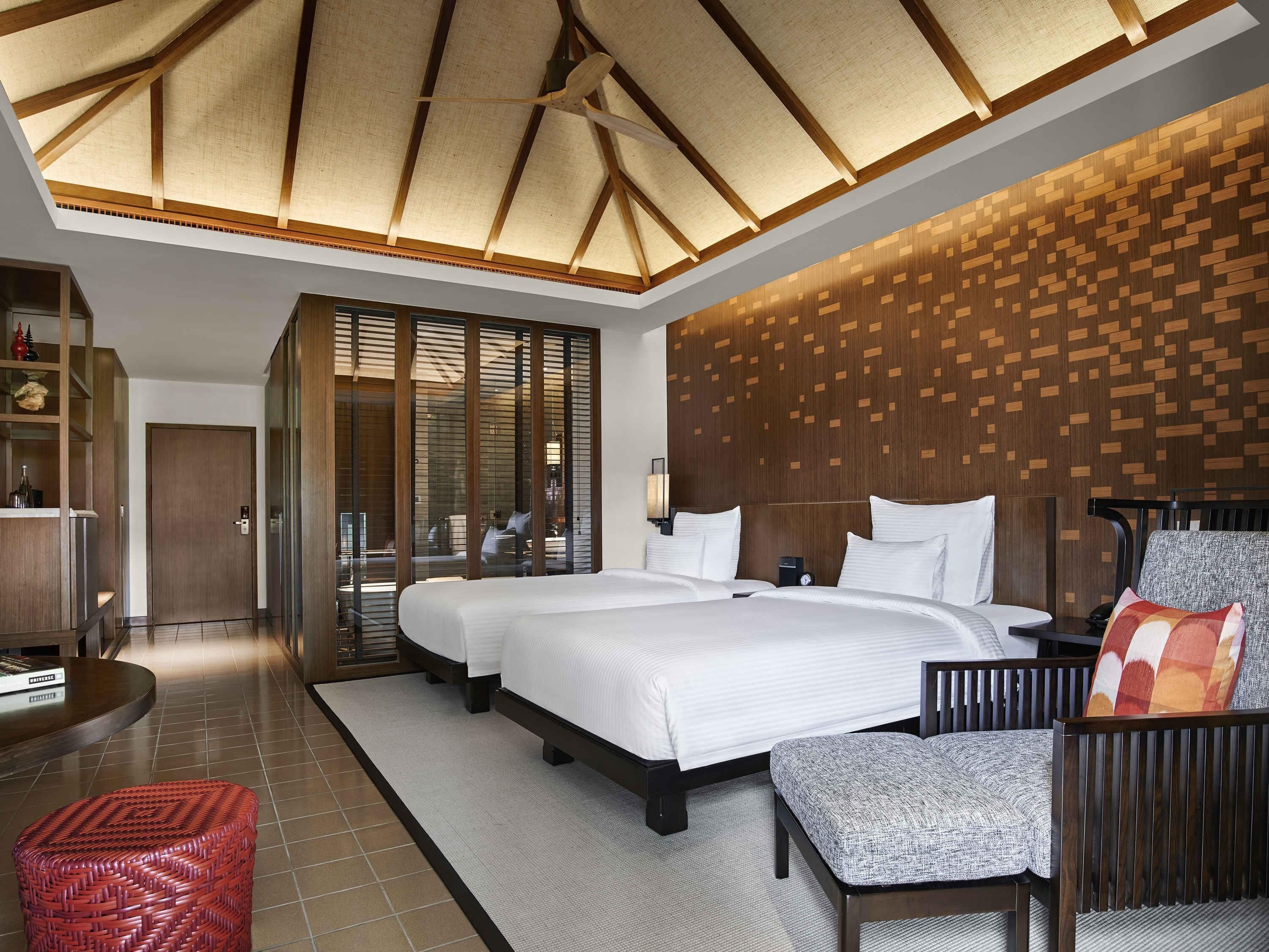 A hotel room with a large bed, a seating area with a chair and ottoman, a wooden wall panel, and a ceiling fan.