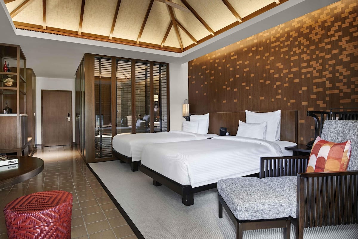 Quarto de hotel com uma cama grande, área de estar com uma poltrona e apoio para os pés, painel de parede de madeira e um ventilador de teto.