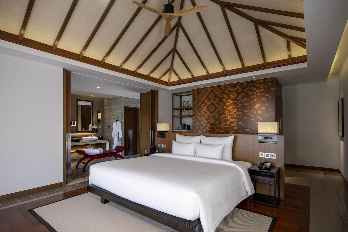 A modern hotel room with a large bed, a ceiling fan, a wooden ceiling, a bathroom with a sink and mirror, and a wall with a patterned design.
