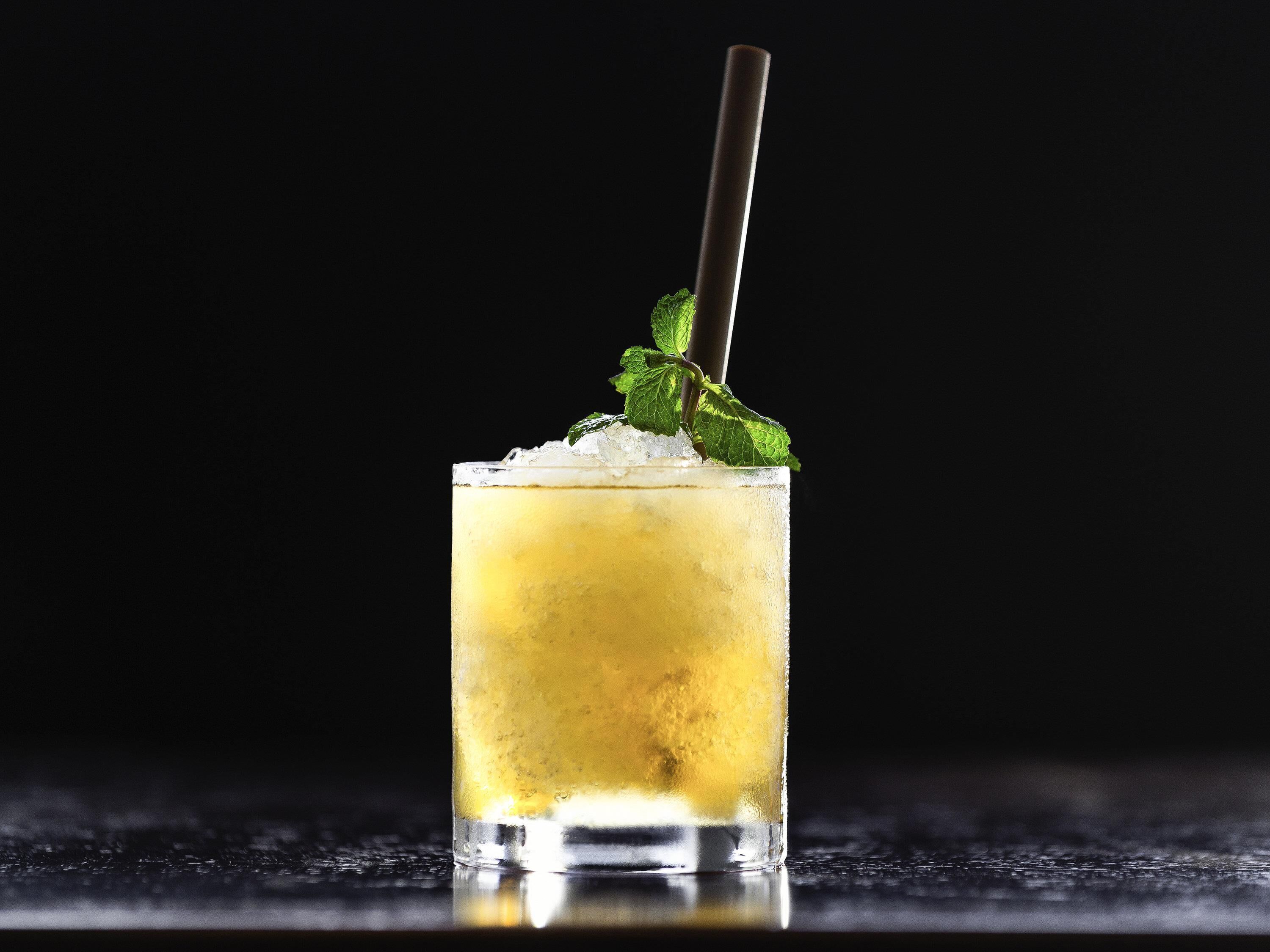 Un vaso alto con pajita y adorno de menta, lleno de una bebida amarilla y cubitos de hielo, sobre un fondo oscuro.