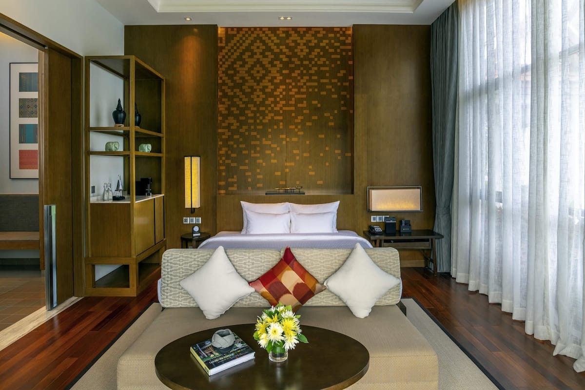 A hotel room with a large bed, a bedside table with a lamp, a nightstand with a phone, a bookshelf, and a view of the city through sheer curtains.