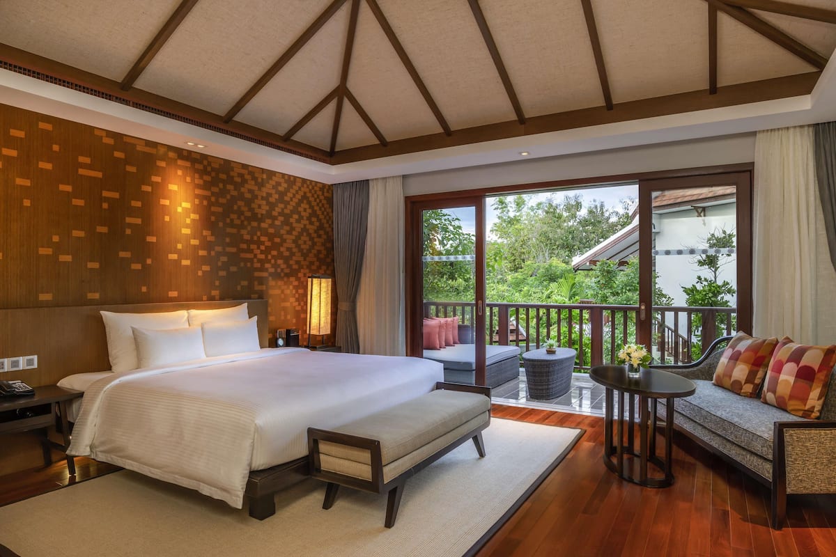 Habitación de hotel con una cama grande, un sofá, una mesita y un balcón con vistas a zonas verdes.