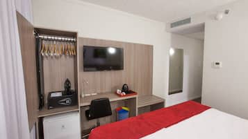 A hotel room with a bed, a desk with a computer, a TV, a mini-fridge, and a coat rack.