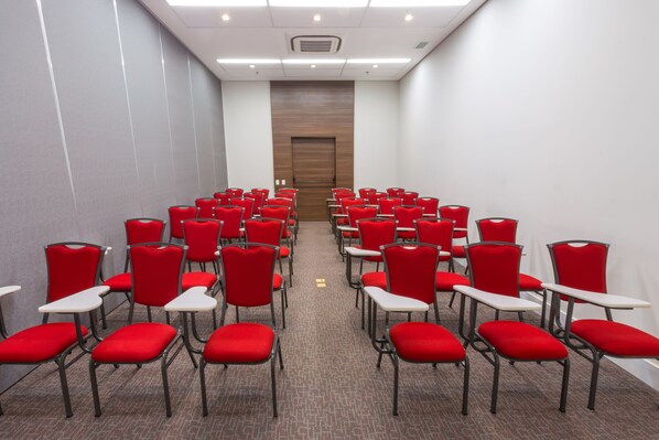 Un salón de conferencias con filas de sillas rojas y mesas blancas, una puerta de madera al fondo, y piso alfombrado.