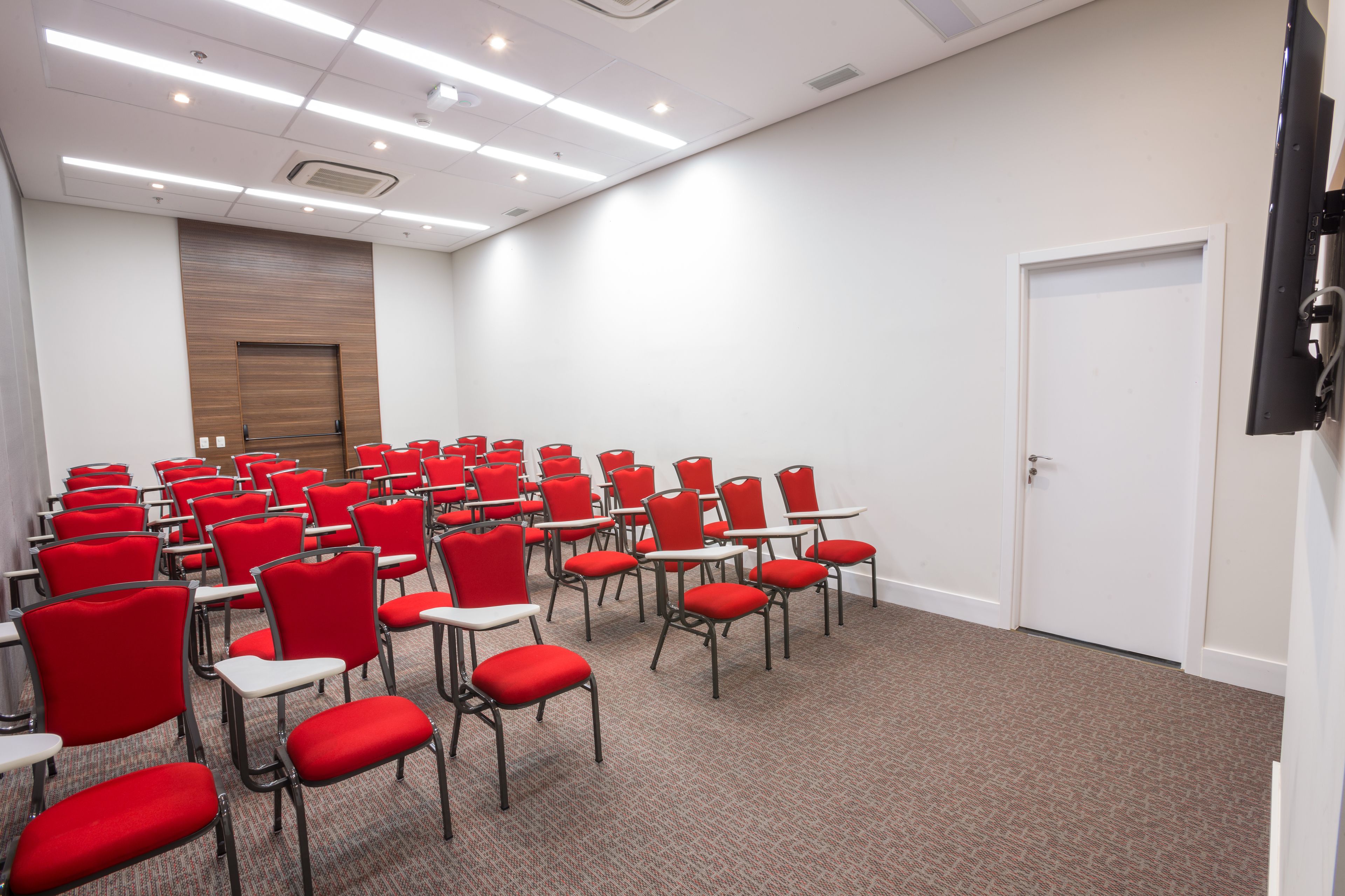 A conference room with rows of red chairs and white tables, a wooden panel wall, and a white door.