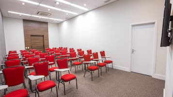 Uma sala de conferências com fileiras de cadeiras vermelhas e mesas brancas, uma parede revestida de painéis de madeira e uma porta branca.