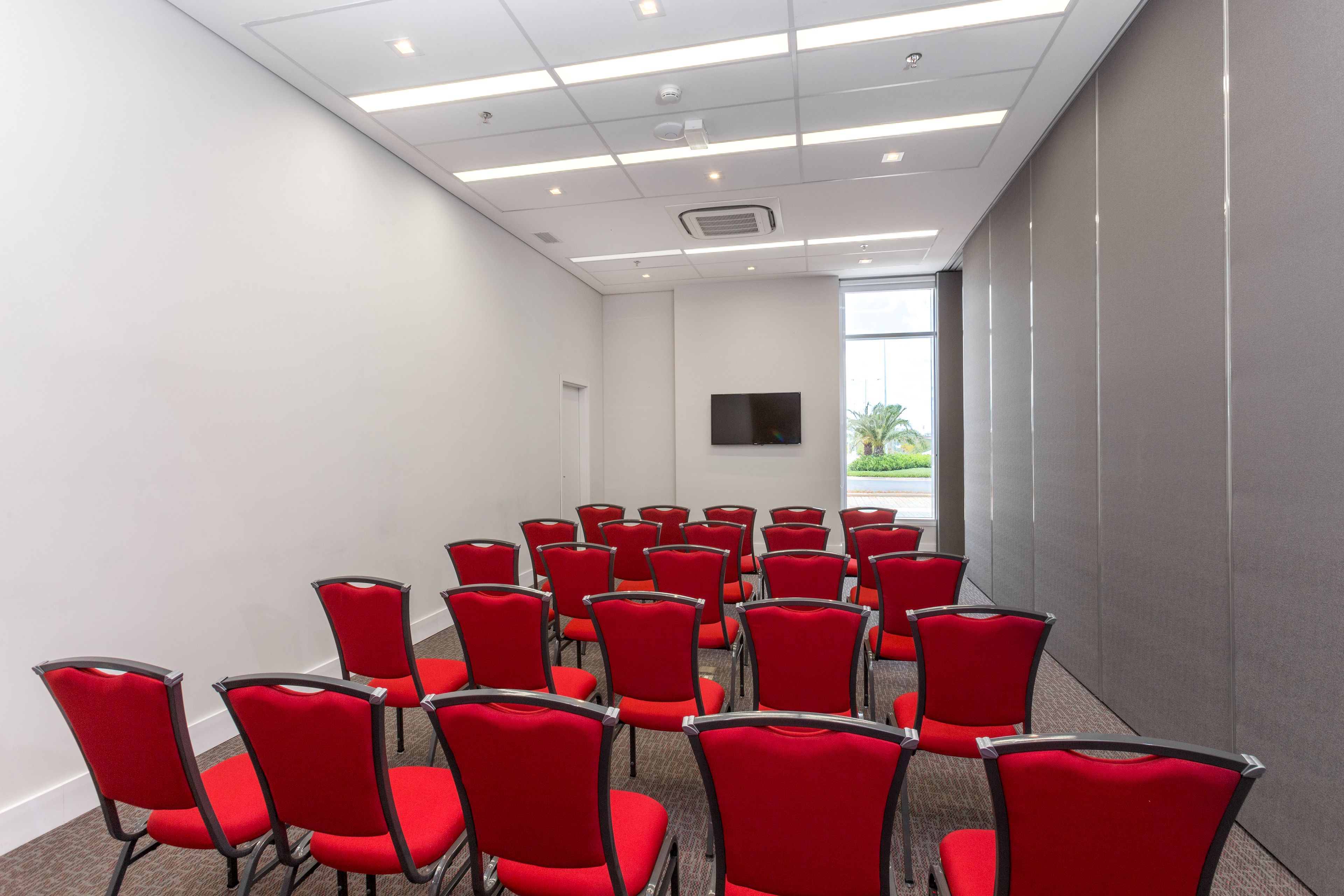 A conference room with rows of red chairs, a flat-screen TV on the wall, and a window with a view of greenery.