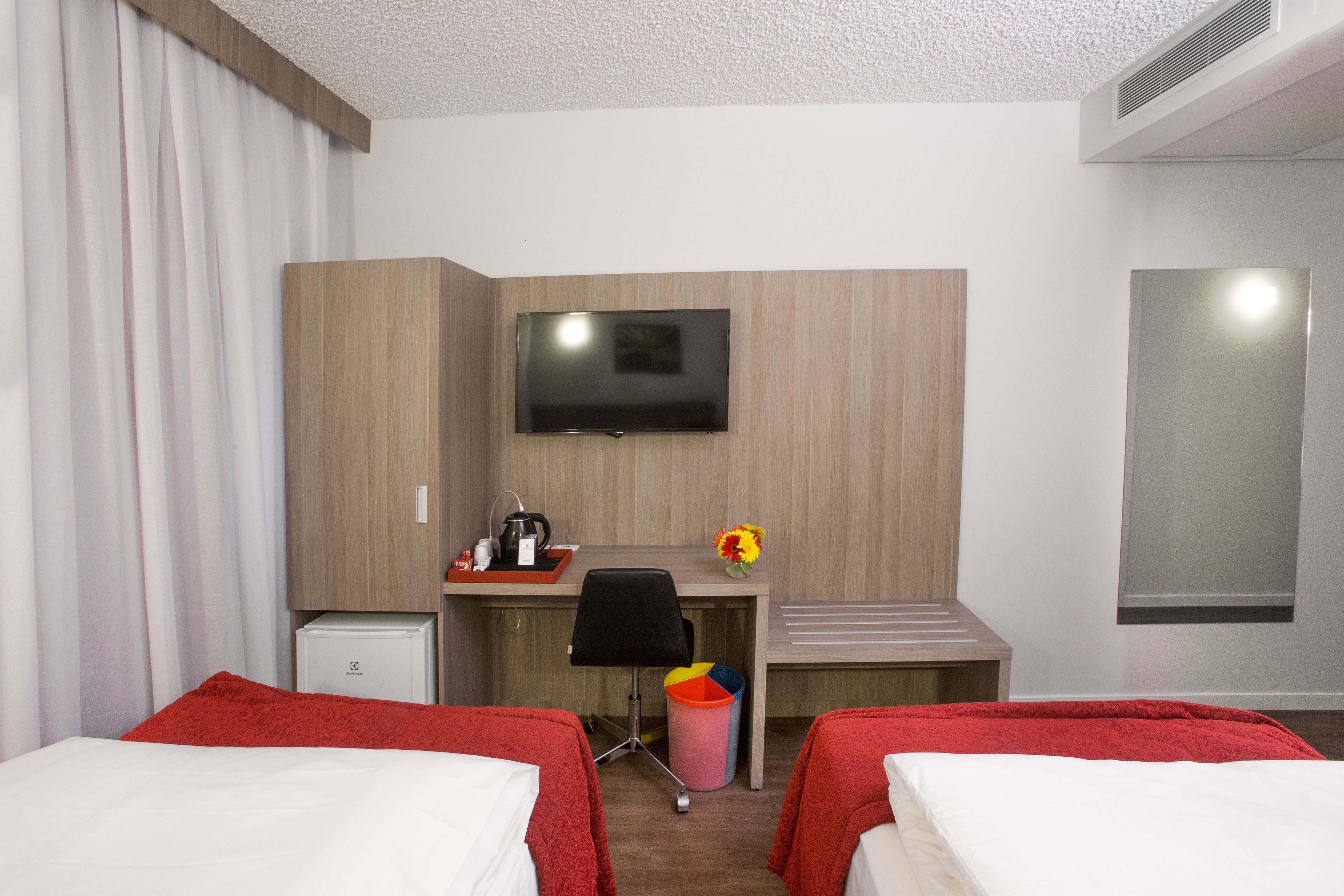 Habitación de hotel con dos camas, un escritorio con televisor, un refrigerador pequeño y un cubo de basura.