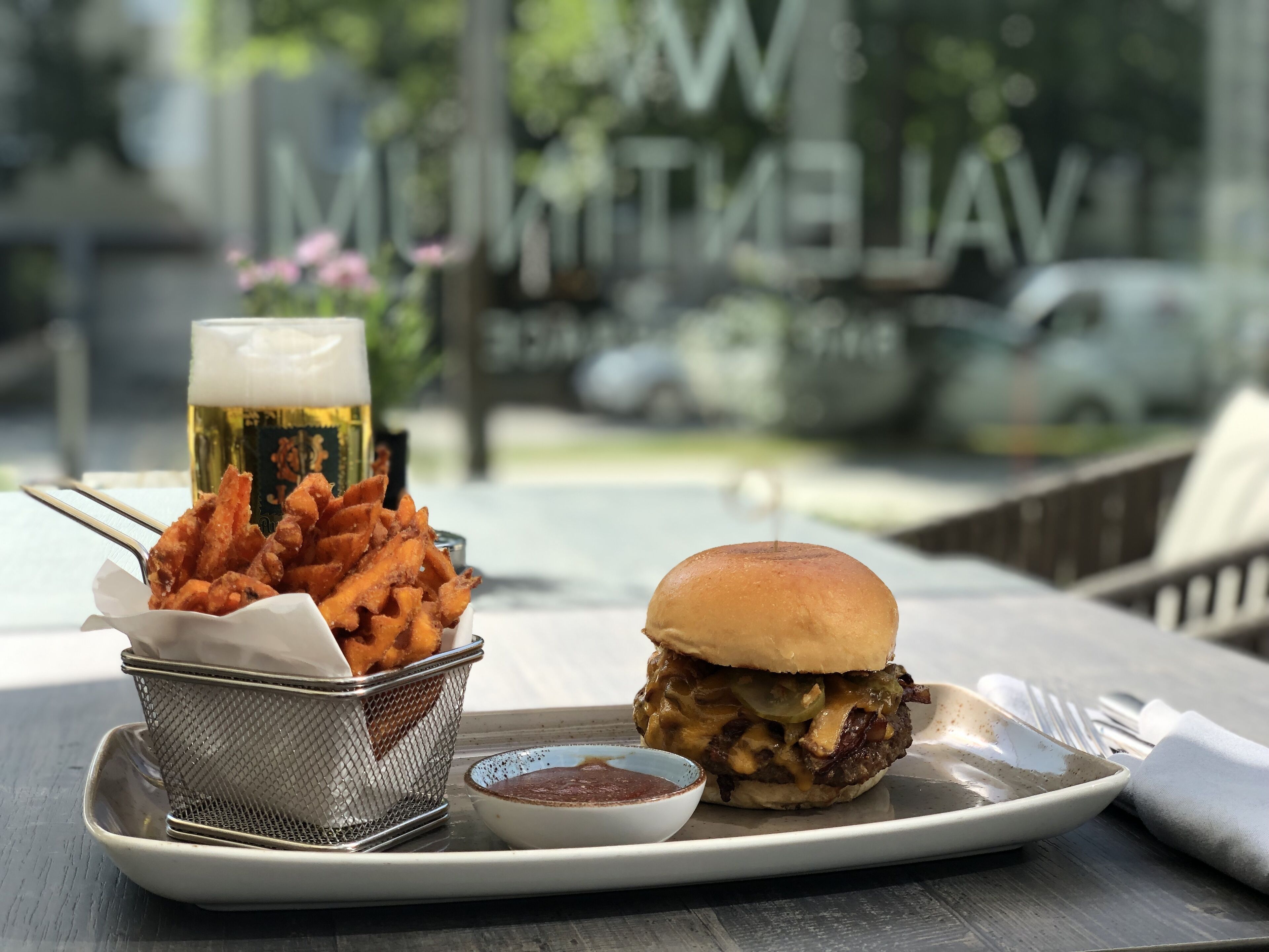 A meal consisting of a burger, fries, and a beer on a table with a view of a street and parked cars.