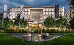 A modern hotel with a glass facade, a fountain in the courtyard, and flags on poles. - Steigenberger Hotel München (Munich)