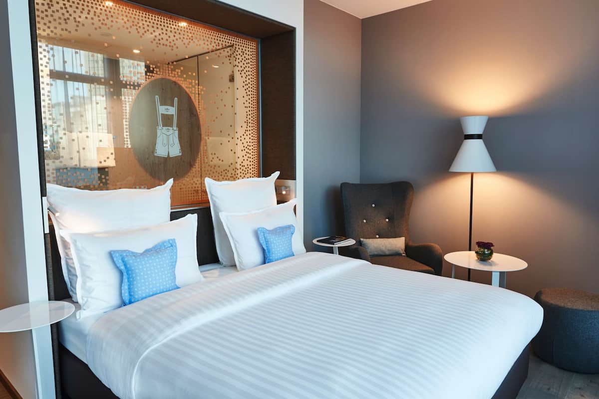 A hotel room with a large bed, a round mirror, a chair, a small table, and a lamp.