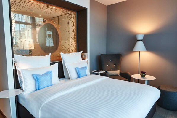 A hotel room with a large bed, a round mirror, a chair, a small table, and a lamp. - Steigenberger Hotel München (Munich)